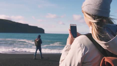 Young-loving-couple-taking-pictures-on-smartphone-on-beautiful-beach-in-Iceland-during-sunset,-slow-motion