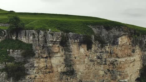 Young-hipster-woman-is-sitting-on-the-edge-on-the-grass-of-tall-wall-of-rock.-Aerial-view.-Drone-is-flying-slow-backward-from-model.-Establishing-revealing-shot