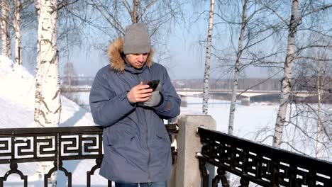 Man-in-blue-down-jacket-with-fur-hood-take-his-cellphone-and-takes-off-the-glove-in-a-winter-Park.