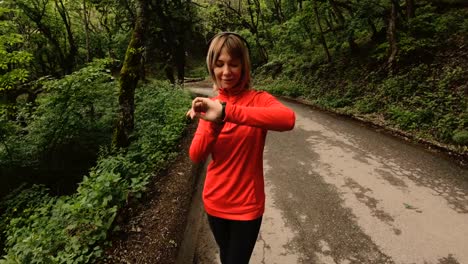 Young-attractive-woman-in-headphones-changing-the-settings-on-a-smart-watch-in-front-of-or-psole-jogging-on-the-road-in-a-green-forest