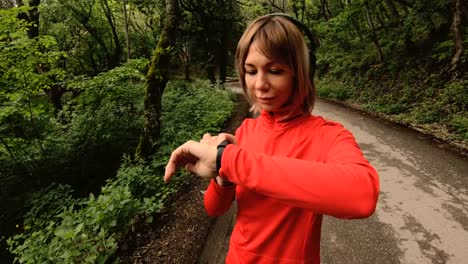 Young-attractive-woman-in-headphones-changing-the-settings-on-a-smart-watch-in-front-of-or-psole-jogging-on-the-road-in-a-green-forest