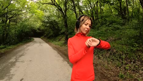 Young-attractive-woman-in-headphones-changing-the-settings-on-a-smart-watch-in-front-of-or-psole-jogging-on-the-road-in-a-green-forest