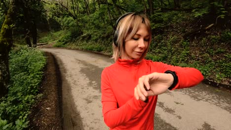 Young-attractive-woman-in-headphones-changing-the-settings-on-a-smart-watch-in-front-of-or-psole-jogging-on-the-road-in-a-green-forest