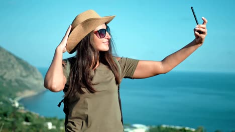 Portrait-of-smiling-woman-traveler-in-hat-and-sunglasses-posing-and-taking-selfie-using-smartphone