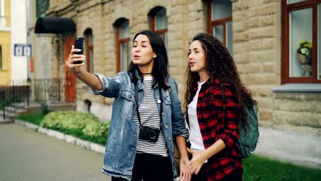 Emotional-young-women-travelers-are-making-online-video-call-using-smartphone-holding-device-and-talking-showing-historical-building-behind-them-expressing-excitement.