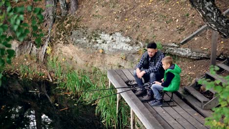 Little-boy-is-fishing-in-lake-with-his-daddy-from-wooden-pier-on-autumn-day,-child-is-holding-rod-sitting-on-chair-while-his-father-is-talking-to-him.-Nature-and-people-concept.