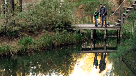 Little-boy-is-learning-to-catch-fish-from-his-father-standing-together-on-wooden-pier-and-talking-holding-fishing-rods.-beautiful-lake-and-forest-are-visible.