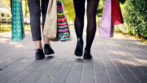 Low-angle-shot-of-women's-legs-walking-in-the-street-with-colorful-paper-bags-after-busy-day-in-shops.-Young-people,-shopping-and-friendship-conccept.