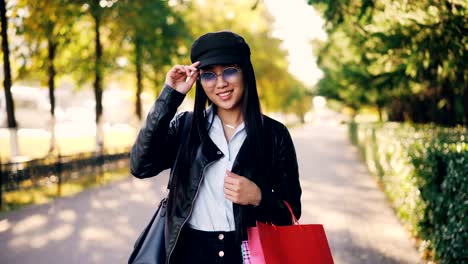 Portrait-of-cute-Asian-girl-in-leather-jacket-and-cap-looking-at-camera,-smiling-and-holding-shopping-paper-bags-standing-outdoors-in-the-street-in-big-city.