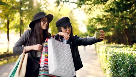 Felizes-mujeres-jóvenes-están-haciendo-videollamada-en-línea-con-teléfono-inteligente-hablar-mostrando-bolsos-de-compras-y-riendo-de-pie-en-la-calle-en-día-de-otoño.