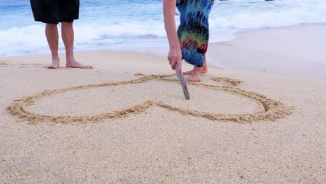 Eine-ältere-Frau-zieht-ein-Herz-und-Initialen-in-den-Sand-und-Ihr-Mann-gibt-ihr-einen-Kuss-zuwerfen