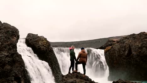 Back-view-of-young-couple-standing-on-a-rock-and-looking-on-a-powerful-waterfall-in-Iceland,-holding-hands