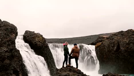 Rückansicht-des-jungen-Paares-auf-einem-Felsen-steht-und-schaut-auf-einen-mächtigen-Wasserfall-in-Island,-Hand-in-Hand