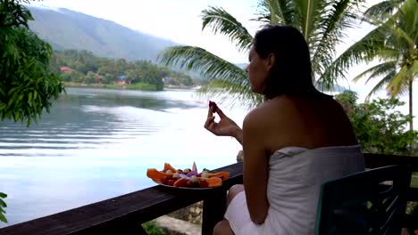 A-woman-sits-in-a-towel-against-the-backdrop-of-a-lake-and-mountains-and-eats-the-Pitahaya