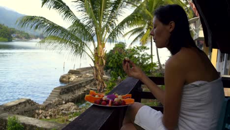 Woman-eating-a-longan-fruit-sitting-in-a-towel-against-the-backdrop-of-a-lake-and-mountains