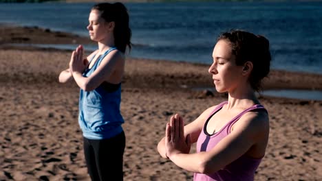 Two-woman-stretching-yoga-standing-on-the-beach-by-the-river-in-the-city.-Beautiful-city-view.-Namaste-pose.