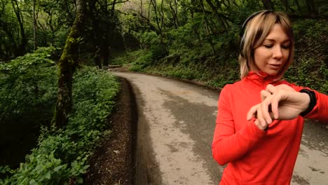 Young-attractive-woman-in-headphones-changing-the-settings-on-a-smart-watch-in-front-of-or-psole-jogging-on-the-road-in-a-green-forest