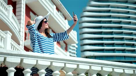 Glamour-woman-in-hat-and-sunglasses-taking-selfie-using-smartphone-standing-on-hotel-terrace