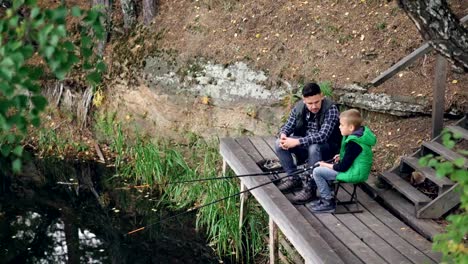 High-angle-view-of-fisherman-and-his-child-serious-boy-fishing-from-wooden-pier-sitting-on-chairs-with-rods-and-talking.-Family,-hobby,-nature-and-relationship-concept.