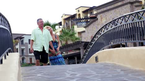 An-older-couple-holding-hands-and-walking-over-a-small-bridge-at-a-resort