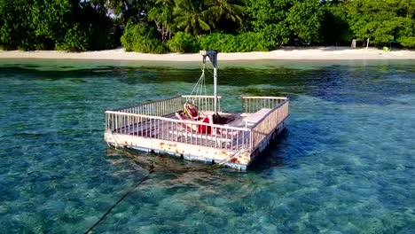 v03857-Aerial-flying-drone-view-of-Maldives-white-sandy-beach-2-people-young-couple-man-woman-relaxing-on-sunny-tropical-paradise-island-with-aqua-blue-sky-sea-water-ocean-4k-floating-pontoon-jetty-sunbathing-together