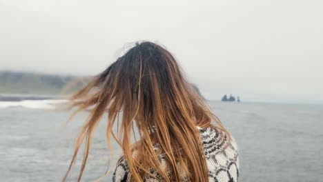 Back-view-of-young-brunette-woman-standing-on-the-black-beach-and-enjoying-beautiful-view-of-troll-toes-rocks-in-Iceland