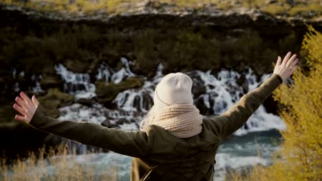 Back-view-of-young-beautiful-woman-standing-near-the-waterfall-and-raising-hands-up,-feeling-freedom-and-happy