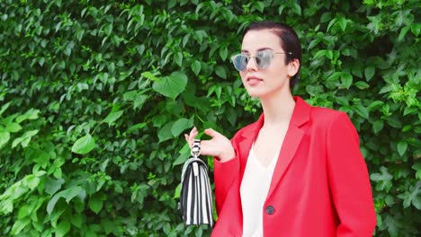 Stylish-Woman-Carrying-Bag-Standing-In-Front-Of-Green-Hedge