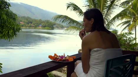 A-woman-sits-in-a-towel-against-the-backdrop-of-a-lake-and-mountains-and-eats-strawberries