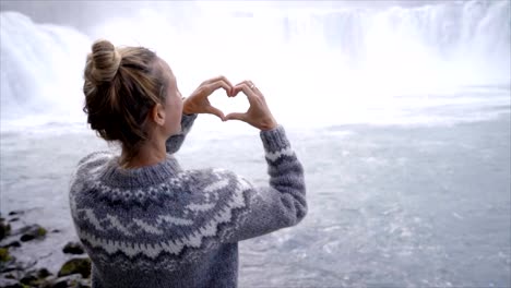 SLOW-MOTION-of-Young-woman-in-Iceland-making-heart-shape-finger-frame-on-spectacular-waterfall