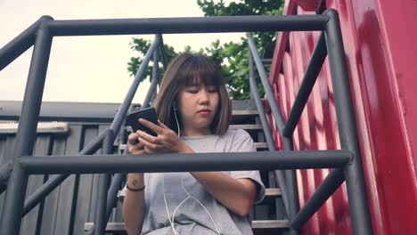 Schöne-junge-asiatische-Frau-anhören-von-Musik-auf-einem-Smartphone-in-der-Stadt.-Junge-asiatische-Frau,-entspannende-Musik-hören-auf-der-Straße.