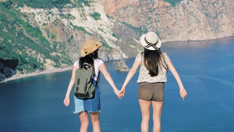 Two-active-elegant-young-traveler-raising-hands-and-having-fun-standing-on-cliff-edge-rear-view