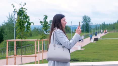 Young-brunette-woman-talking-on-video-chat-on-mobile-phone-walking-along-the-promenade-in-the-city.