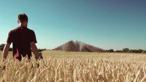 Adult-walking-through-Beautiful-wheat-field-towards-modern-pyramide-holding-skateboard-with-blue-sky-and-epic-sun-light---shot-on-RED