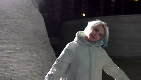 Adult-girl-with-short-dyed-blonde-hair-walking-along-the-street-at-night,-wearing-white-jacket,-ripped-jeans