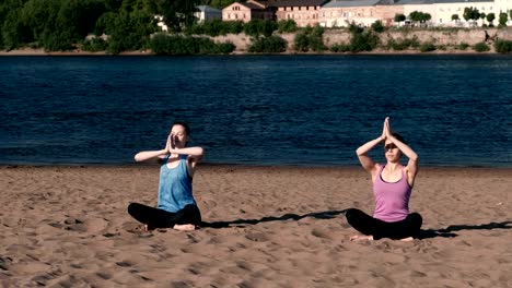 Two-woman-stretching-yoga-on-the-beach-by-the-river-in-the-city.-Beautiful-city-view.-Namaste-pose.