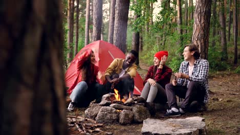 Happy-girls-and-guys-friends-are-talking,-gesturing-and-laughing-resting-around-fire-getting-warm-at-campsite.-Conversation,-nature-and-people-concept.