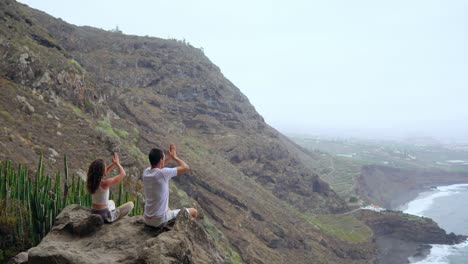 Un-hombre-y-una-mujer-sentada-en-la-cima-de-una-montaña-mirando-el-mar-sentado-en-una-piedra-meditando-en-posición-de-loto.-La-vista-desde-la-parte-posterior.-Islas-Canarias