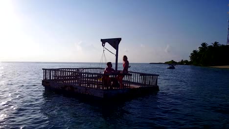 v03828-Aerial-flying-drone-view-of-Maldives-white-sandy-beach-2-people-young-couple-man-woman-relaxing-on-sunny-tropical-paradise-island-with-aqua-blue-sky-sea-water-ocean-4k-floating-pontoon-jetty-sunbathing-together