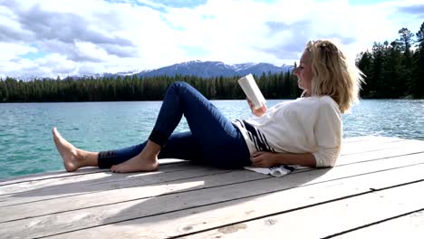 Mujer-joven-relajante-en-el-muelle-del-lago-con-el-libro,-Canadá