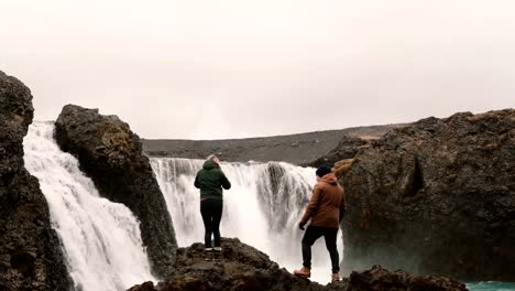 Young-happy-couple-hiking-together-to-see-a-powerful-waterfall-in-Iceland.-Man-and-woman-raising-hands-up