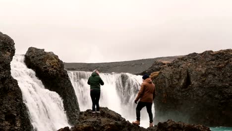 Glückliches-junges-Paar-Wandern-zusammen,-um-einen-mächtigen-Wasserfall-in-Island-zu-sehen.-Mann-und-Frau,-die-Hände-hob