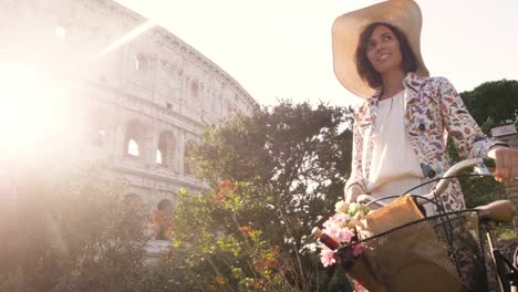 Beautiful-young-woman-in-colorful-fashion-dress-walking-alone-on-hill-with-bike-in-front-of-colosseum-in-Rome-at-sunset-with-trees-attractive-girl-with-straw-hat