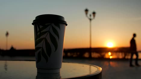 one-coffee-cup-is-standing-on-a-table-of-a-street-cafe-in-background-of-embankment,-sunset-sky