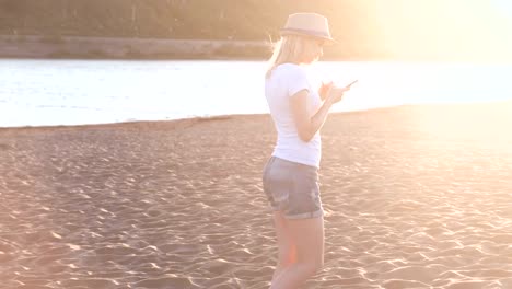 Beautiful-blonde-girl-in-a-hat-types-a-message-on-her-mobile-phone-walking-on-the-beach-at-sunset.