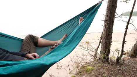 Young-caucasian-male-resting-lying-on-blue-hammock-outdoors-near-lake.