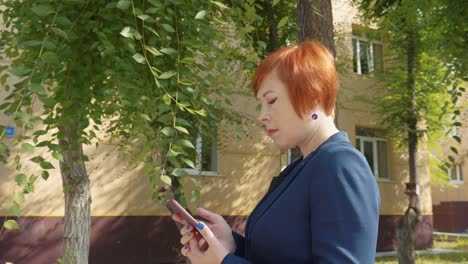 Mujer-pelirroja-atractiva-navegación-teléfono-y-mirando-lejos,-lateral-Vista.-Bonita-mujer-adulta-con-smartphone-al-aire-libre