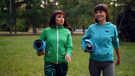 Sonriente-mujer-joven-está-caminando-en-el-parque-con-esteras-de-la-yoga-y-hablando-de-compartirlas-noticias-y-discutían-práctica.-Juventud,-actividad-al-aire-libre-y-el-concepto-de-conversación.