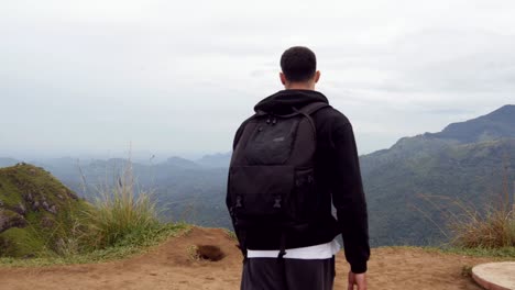 Male-hiker-reaching-up-top-of-mountain-and-outstretching-arms-up.-Young-man-tourist-with-backpack-standing-on-the-edge-of-beautiful-canyon-and-victoriously-raised-hands.-Rear-back-view