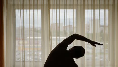 Silueta-de-un-hombre-de-realización-de-ejercicio-en-el-interior.-En-la-ciudad-de-fondo-calle-detrás-de-cortinas-de-la-ventana.-Hacen-los-hombres-calentando-la-mañana-estiramientos.